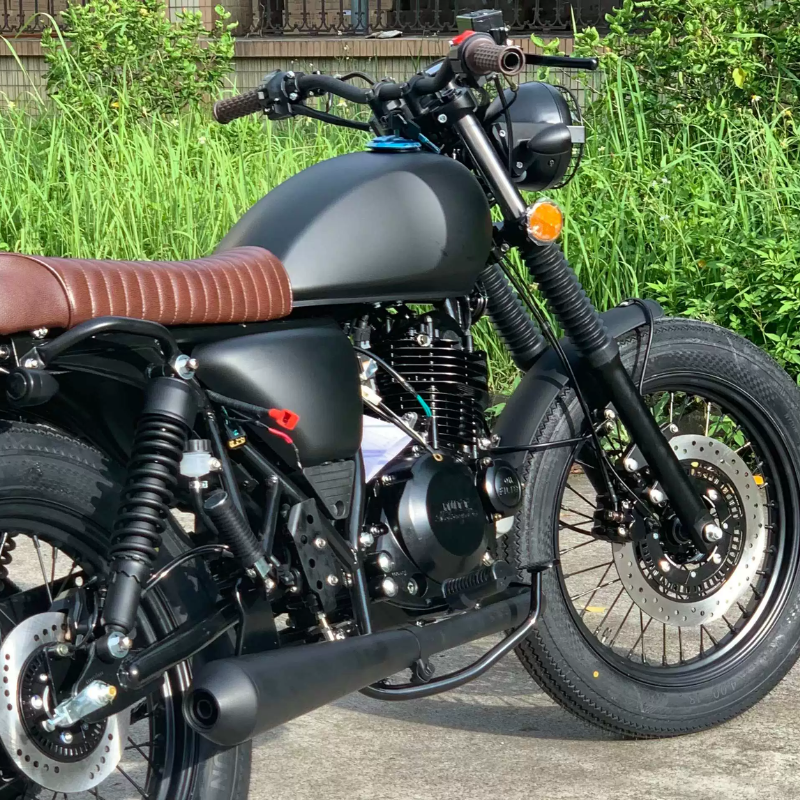 A custom cafe racer motorcycle with a matte black fuel tank, black forks and exhaust, a brown leather seat, and a black spoked wheel, photographed outdoors on a concrete surface with green grass in the background.